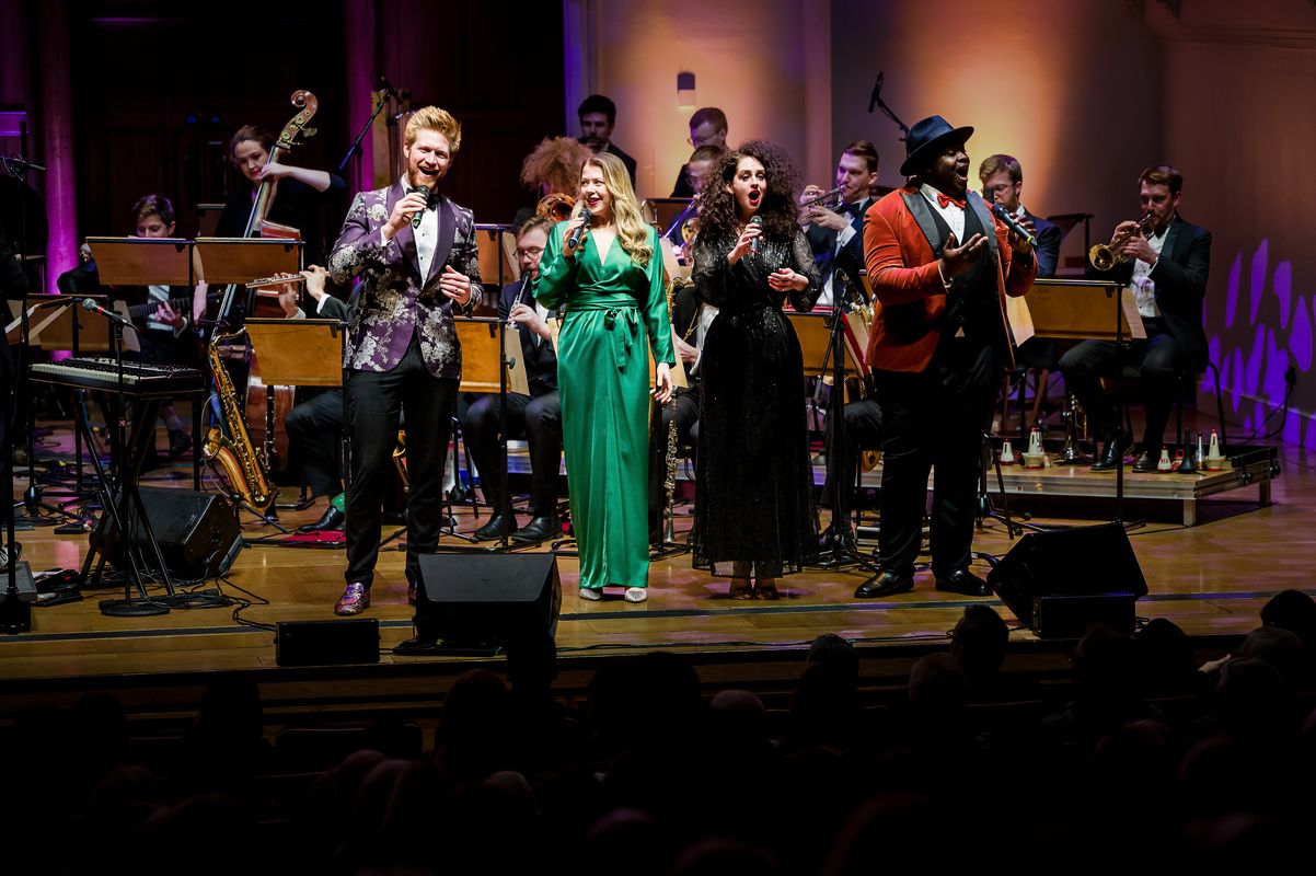 Four people sing on stage with the band playing in the background.