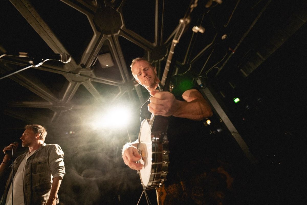 A person playing a banjo on stage, illuminated by bright stage lights. Another person stands nearby holding a microphone. The setting has a geometric ceiling design and a dimly lit atmosphere.