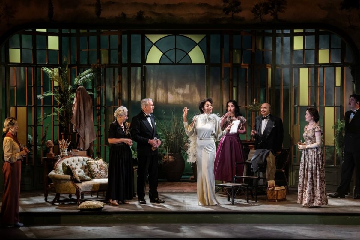 A group of elegant individuals stands on a vintage-styled stage set resembling an ornate conservatory, with decorative furniture and lush plant where actors are dressed in formal evening attire. The setting includes large, arched windows and a misty ambiance, suggesting a classic, sophisticated atmosphere.