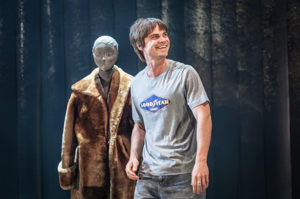 A man is stood smiling and behind him is a mannequin wearing a large brown coat