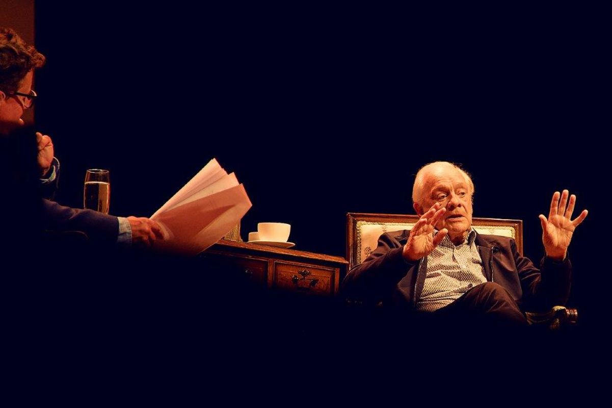 Sir David Jason on stage with another gentleman. The setting is made of two chairs, a dark wooden desk and a large rug