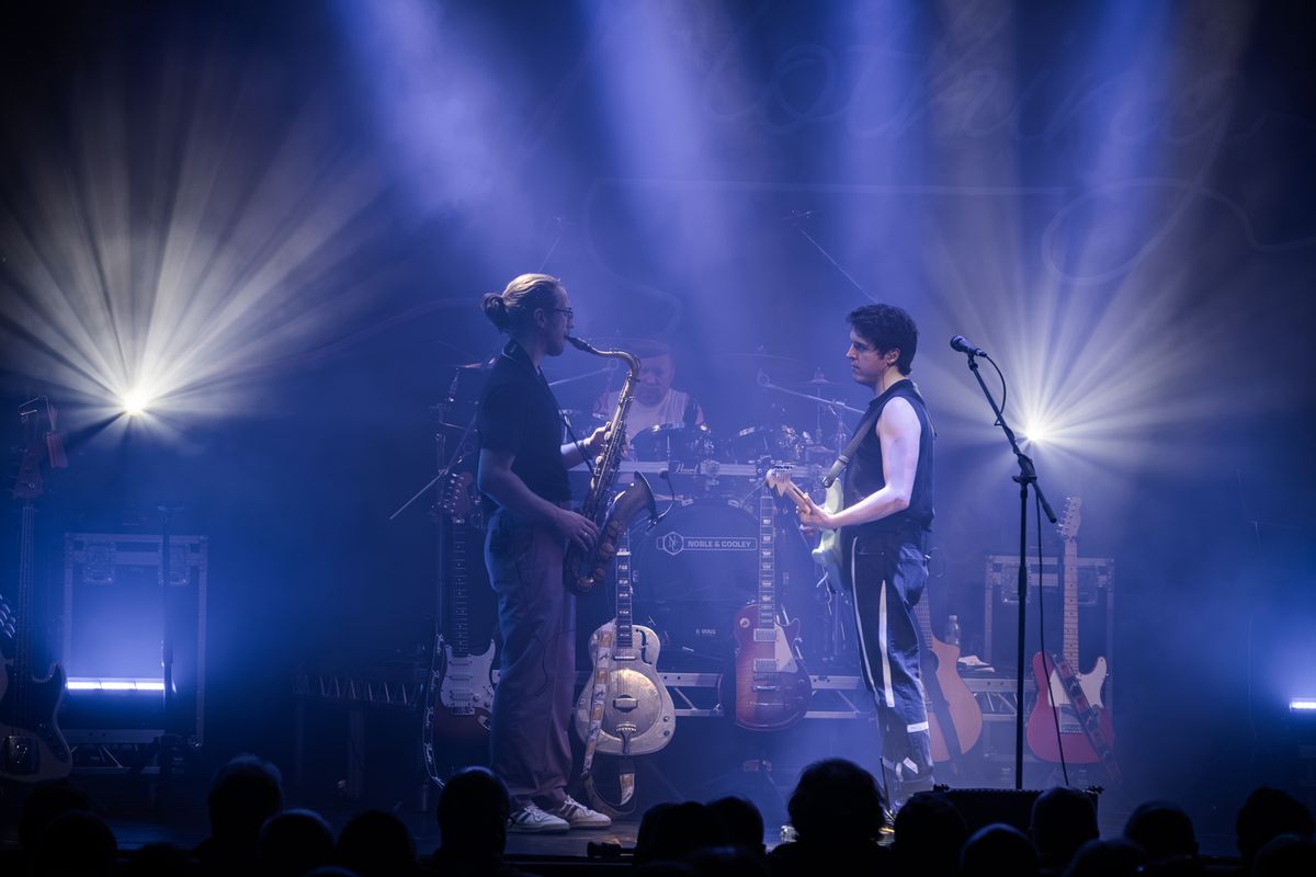 Band on stage performing, they're playing guitars and drums and there are flashing lights around them