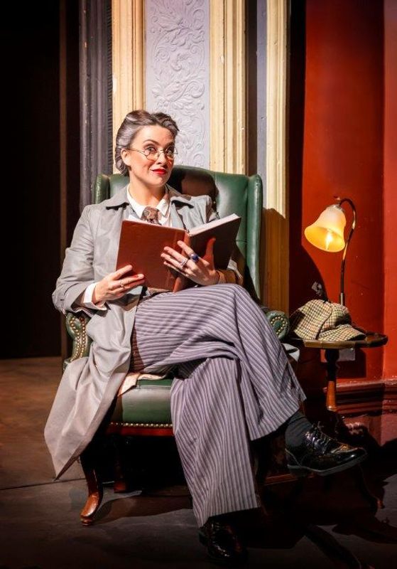 A person with glasses and gray hair sits in a green armchair, reading a book. They wear a gray coat and striped pants. A warm lamp and handbag are beside them, creating a cozy, contemplative atmosphere.