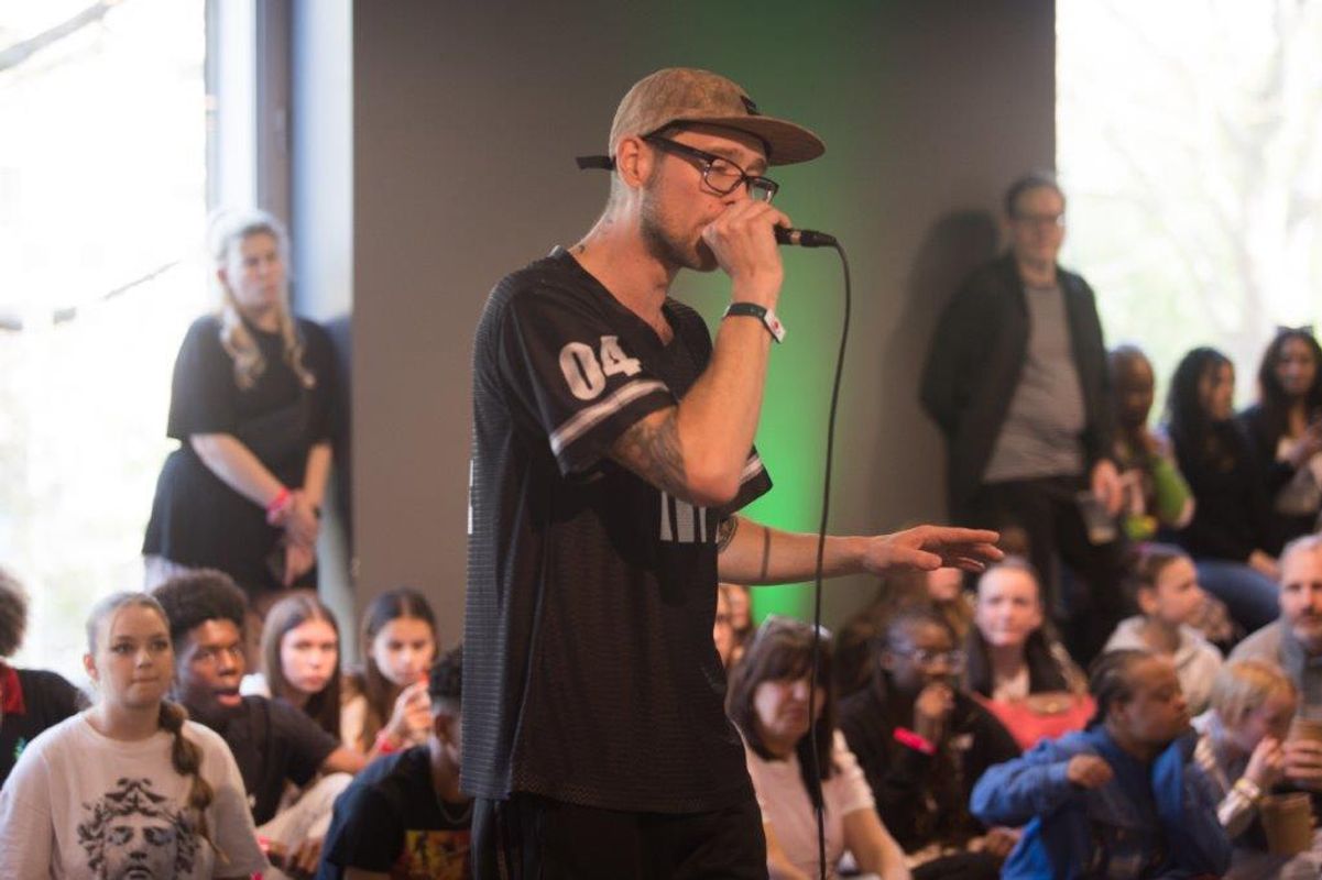 A person wearing a cap and glasses holds a microphone surrounded by a group of young people sat down.