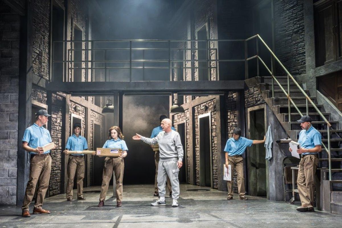There is a group of people stood in a prison setting, they are all wearing beige trousers and light blue polo shirts. There is a man stood in the centre in a grey tracksuit
