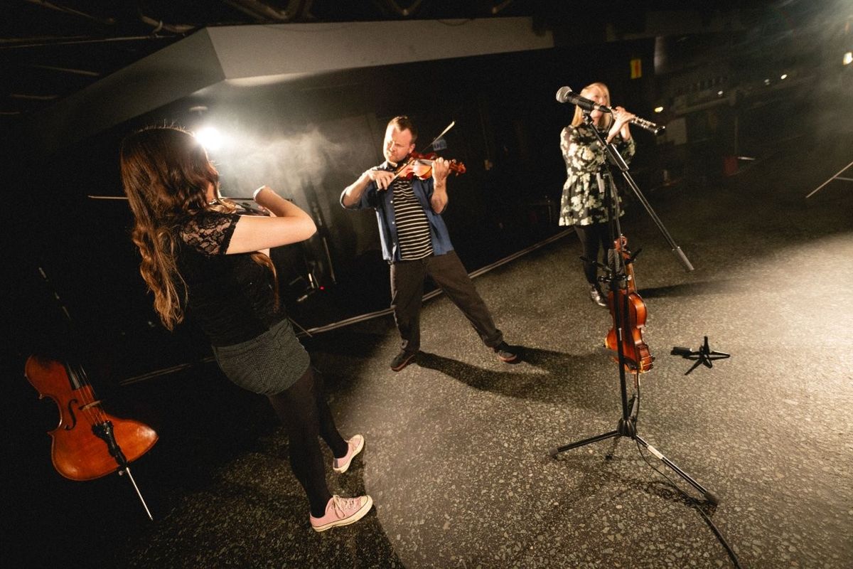 A group of musicians performing on a stage. One person is playing a violin passionately, another is playing a flute, and a cello rests nearby. They are surrounded by dramatic stage lighting.