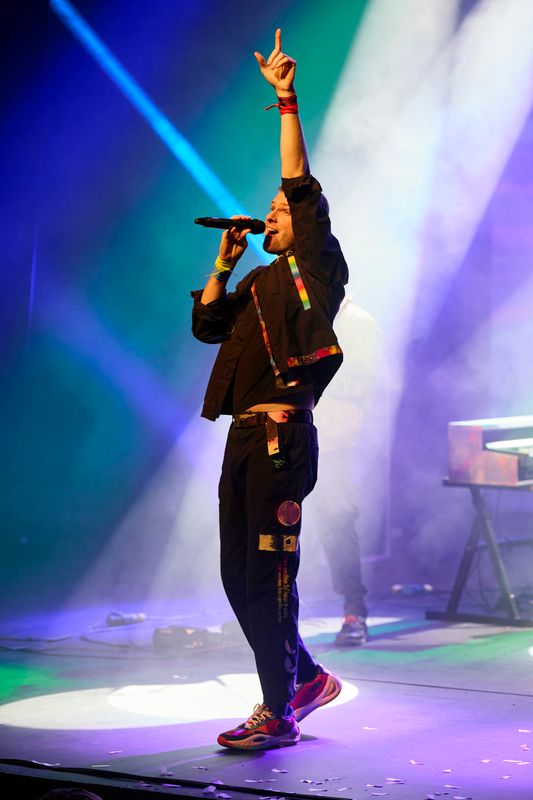 Singer on stage, wearing a colorful outfit, passionately performs under vibrant blue and purple lights. One arm raised, creating an energetic atmosphere.