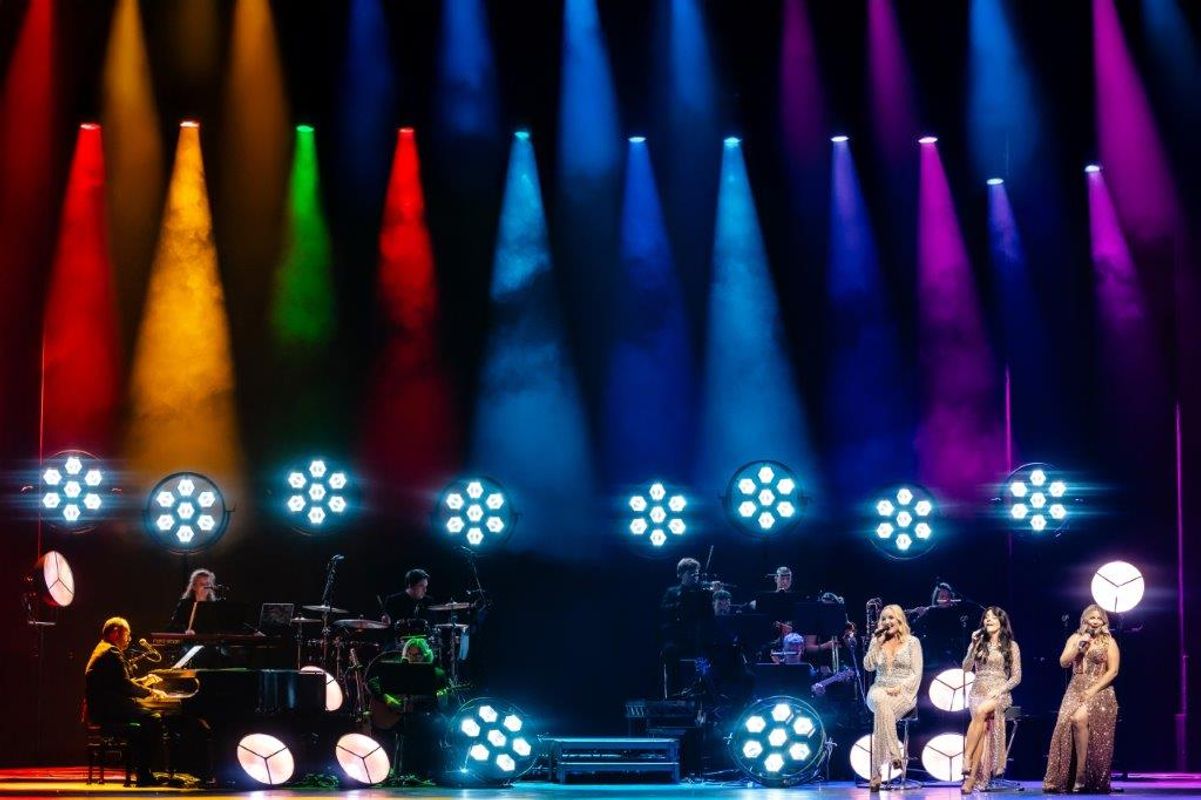 A vibrant concert scene with colorful spotlights in orange, green, blue, and purple. Kerry Ellis, Louise Dearman and Rachel Tucker in sparkling gowns perform with a band and pianist.