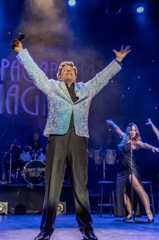Martyn James in a sparkling jacket raises arms triumphantly on stage, microphone in hand. Background dancer strikes a pose. Energetic concert scene.