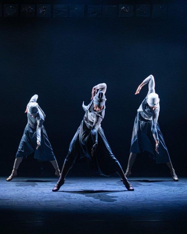 Group of dancers on stage wearing blue and grey outfits, a light casts a shadow over them