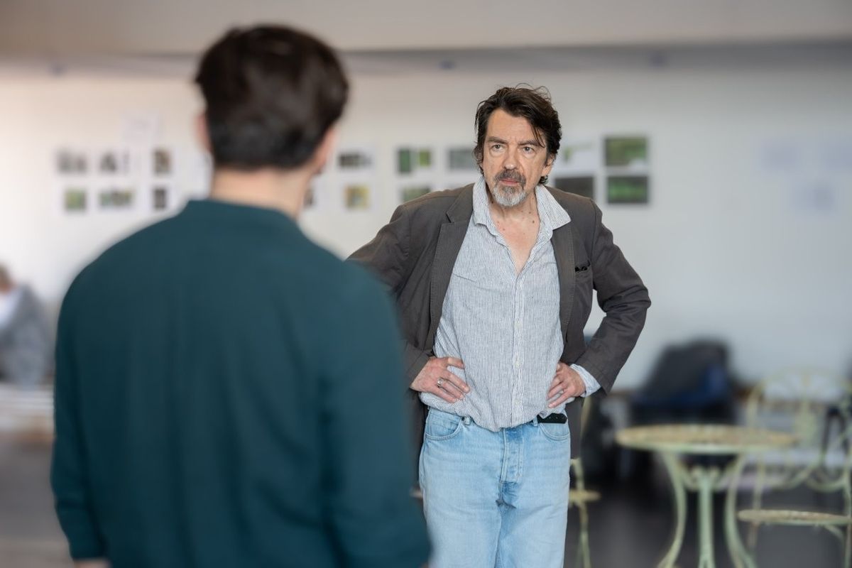 Two men are looking at each other, one has his back to the camera and the other is facing him with his hands on his hips