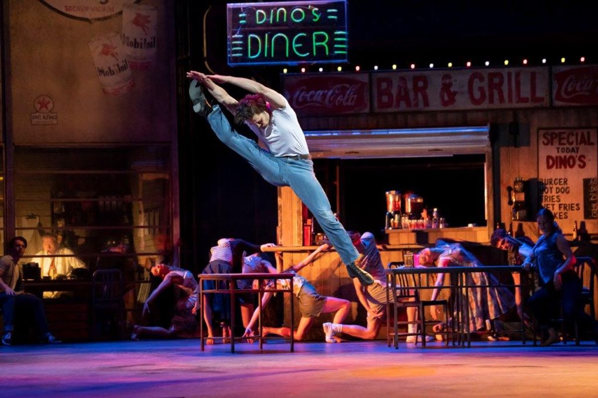 A dancer in jeans and a white shirt leaps energetically on stage at Dino's Diner, with neon lights and performers posing dynamically in the background.
