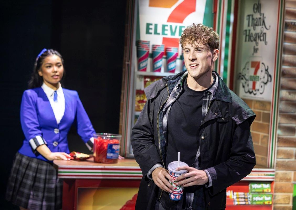 A man in a dark coat sings passionately while holding a 7-Eleven cup on stage. A woman in a blue uniform watches attentively from a counter. Stage lighting creates a dramatic ambiance.