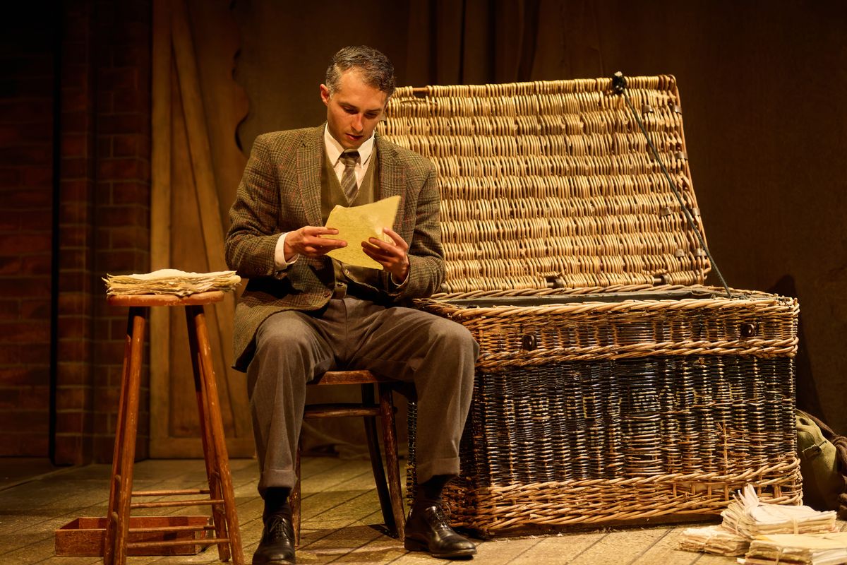 A person sits next to a large wicker chest reading a letter.