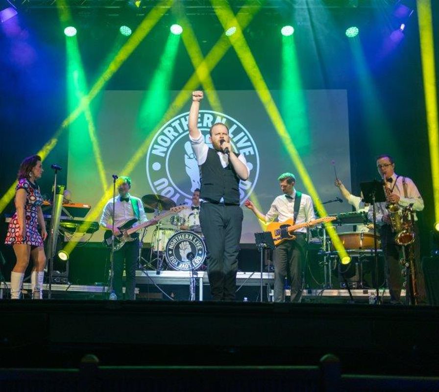 A person dressed in a black waistcoat stands centre stage with their fist in the air.