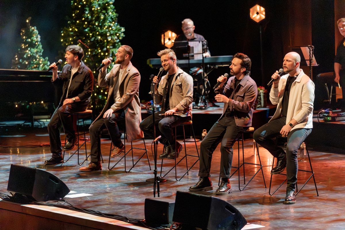 Five members are sat on tall stools singing into microphones