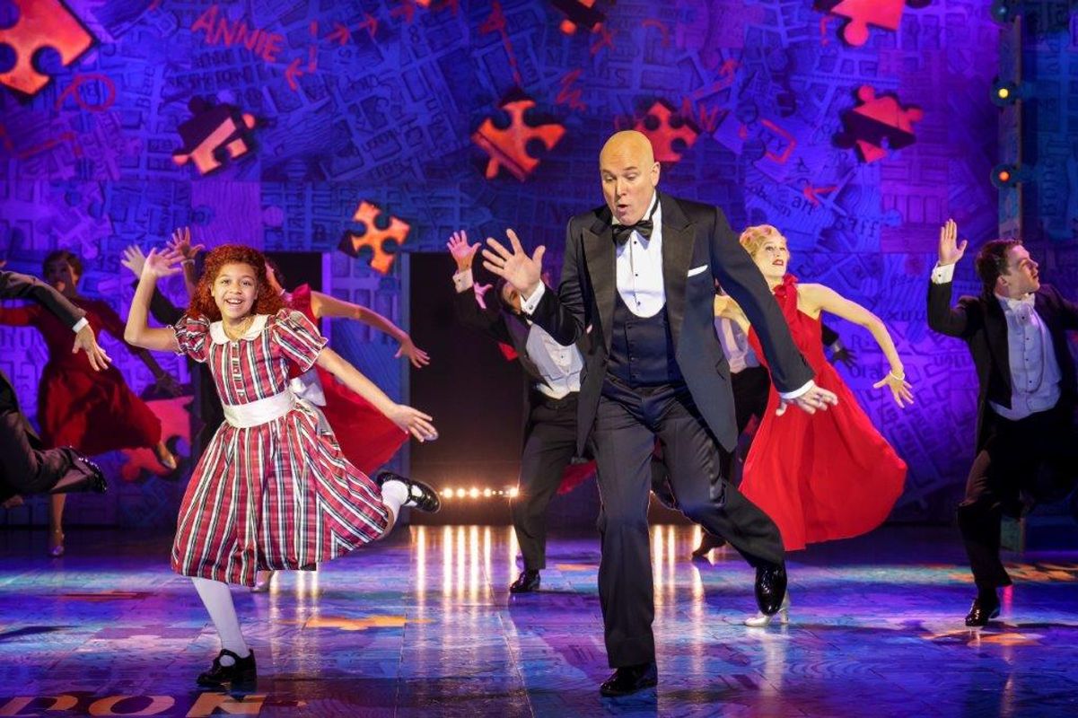 A joyous stage performance with a girl in a plaid dress dancing beside a bald man in a tuxedo. Vibrant background and other dancers enhance the lively atmosphere.