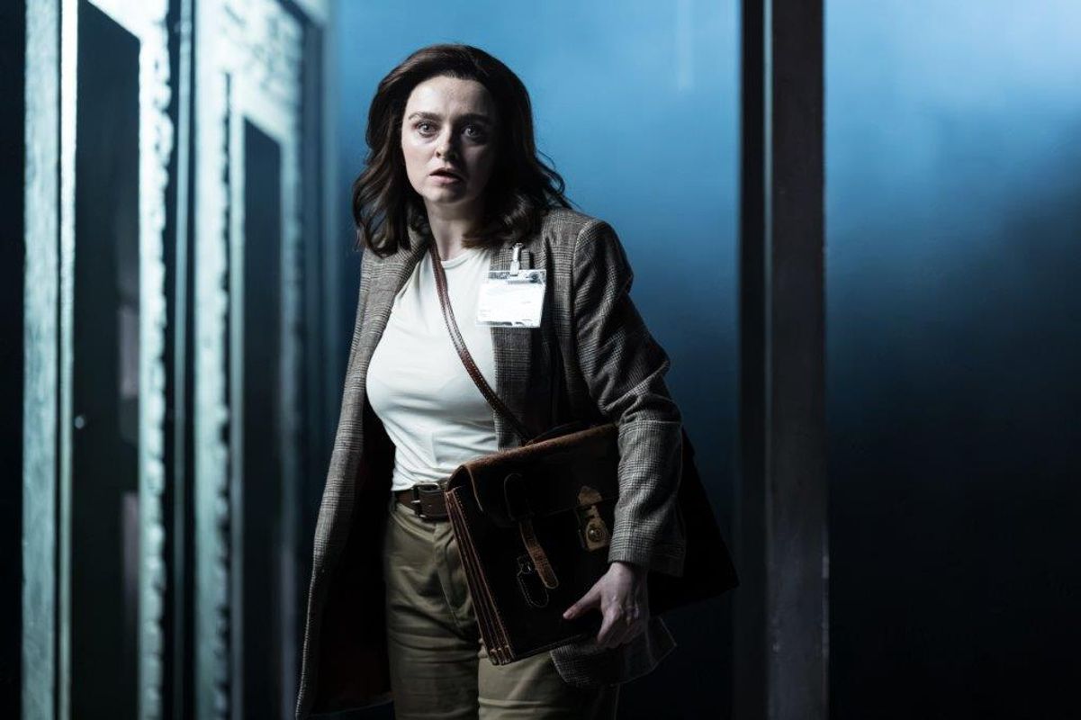 A woman is holding a folder and is wearing a badge, she faces head on
