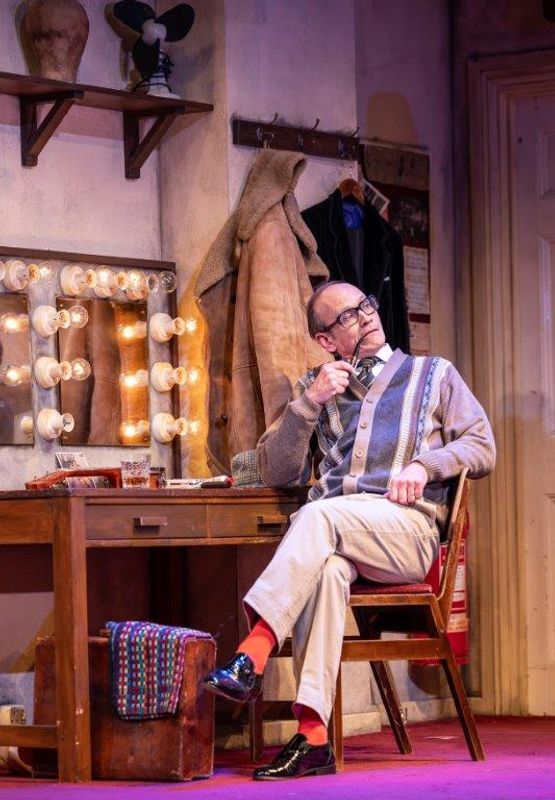 A man wearing glasses and smoking a pipe is sat in a dressing room backstage setting