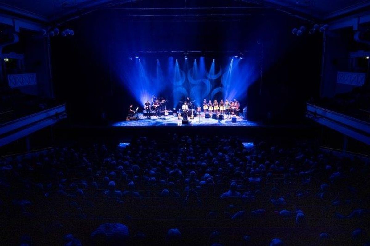 The South African Cultural Choir are singing on stage with Paul Simon. They are all wearing bright and vibrant African clothing. Paul is playing a guitar