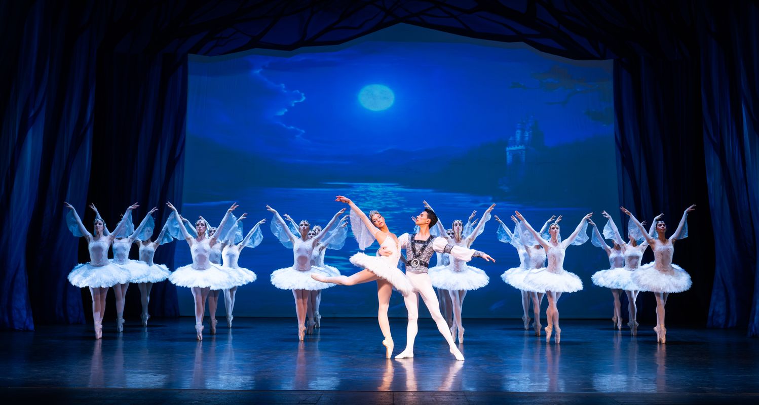 An ensemble of ballet dancers stand on pointe with their arms in the air.