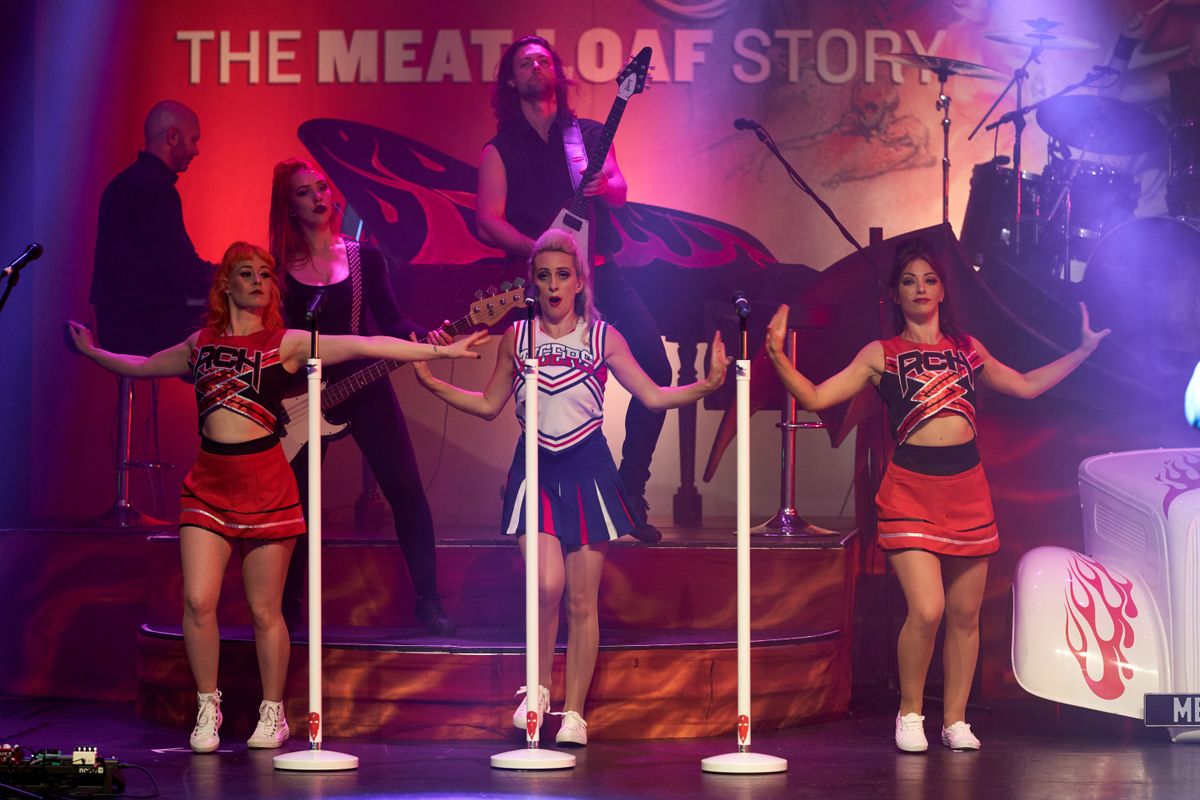 Three women in cheerleader outfits dancing with microphones. Musicians play instruments in the background, creating a vibrant, energetic atmosphere.