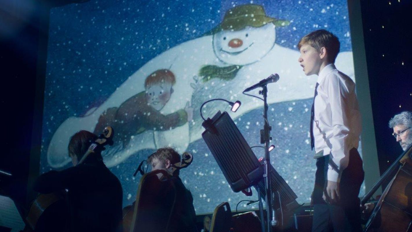 People performing on stage with The Snowman on a projector behind them.