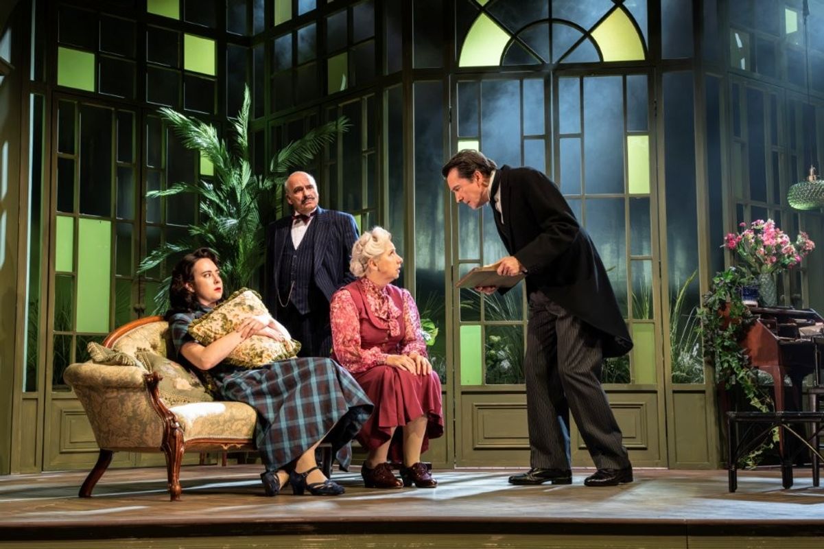 A theatrical scene depicting four performers on stage in a set resembling a lush, elegant room with large windows. One performer holds a cushion while seated on a vintage sofa beside another onlooker dressed in a formal suit. Two others are standing, with one leaning forward in conversation. The background includes potted plants and period-style decor, contributing to the vintage atmosphere.
