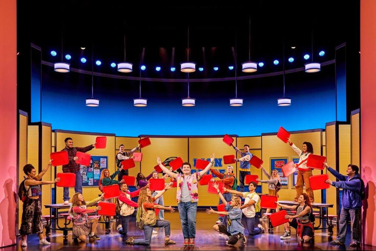 Stage performance with a lively group of people holding red chairs above their heads. The colorful set and bright lighting create an energetic atmosphere.
