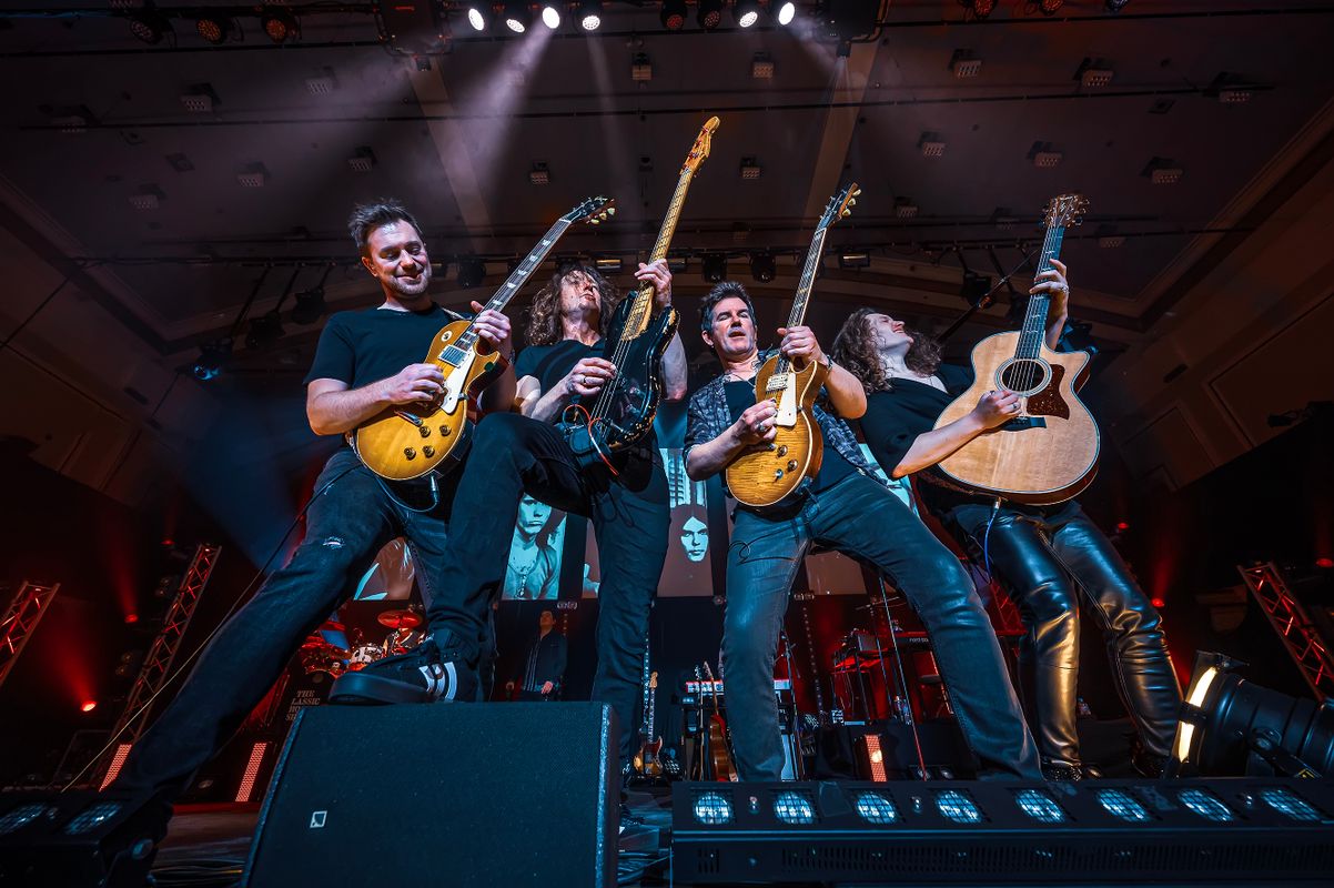 Four people stand in a line next to each other playing guitar.