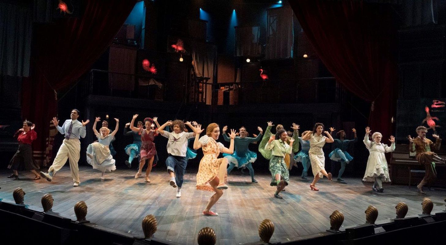 A larger number of people dancing on stage, all have their hands and foot up.