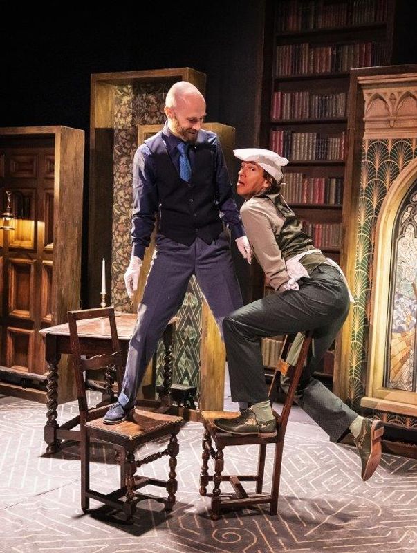 Two performers stand on chairs in a playful, theatrical scene. One wears a vest and gloves, the other a hat and suspenders. Shelves of books are in the background.