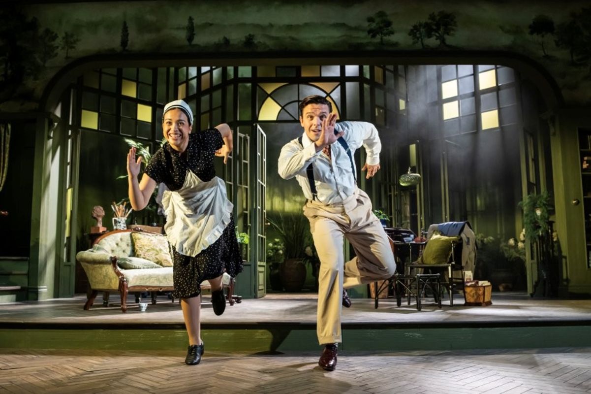 Two performers are energetically dancing onstage in front of a theatrical set resembling a living room. They appear joyful, wearing vintage-style clothing. The set includes a sofa and a lamp.