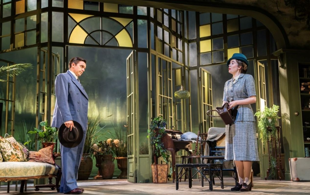 A theatrical scene set in a conservatory with arched windows, showcasing two people. One is holding a hat, and the other is dressed in a suit with a hat, holding a watering can. The stage is decorated with plants and garden furniture.