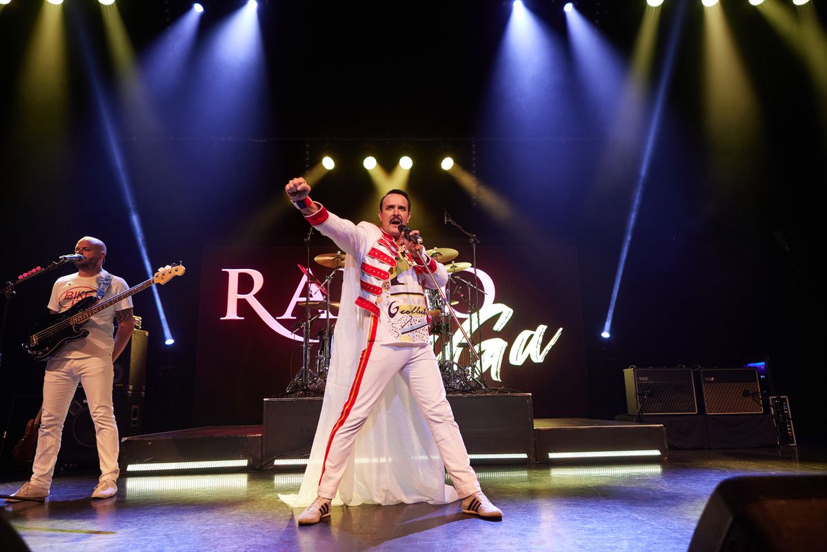 Performers singing and playing instruments on stage to an audience, bright lights are shining on the band