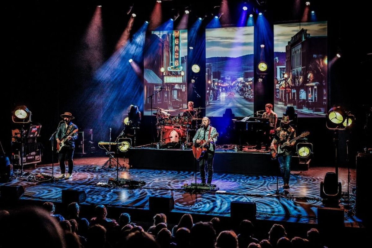 The image shows a band performing on stage with several musicians playing guitars, keyboards, and drums. The backdrop features a vibrant street scene with colorful lights, resembling a theater district or cityscape. The stage is illuminated with dynamic spotlights. An audience is visible in the foreground.