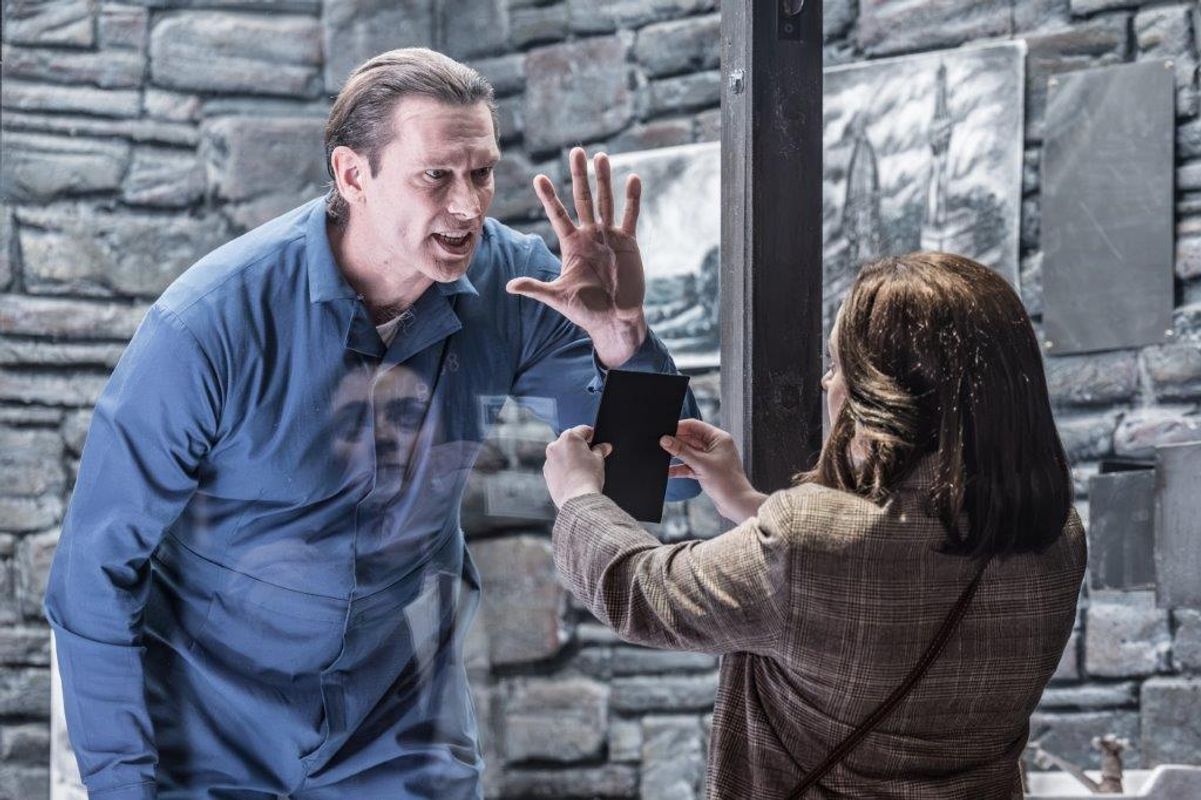 Two people facing each other, one is behind a glass cell, the other is showing him her badge, he has his hand pressed against the glass