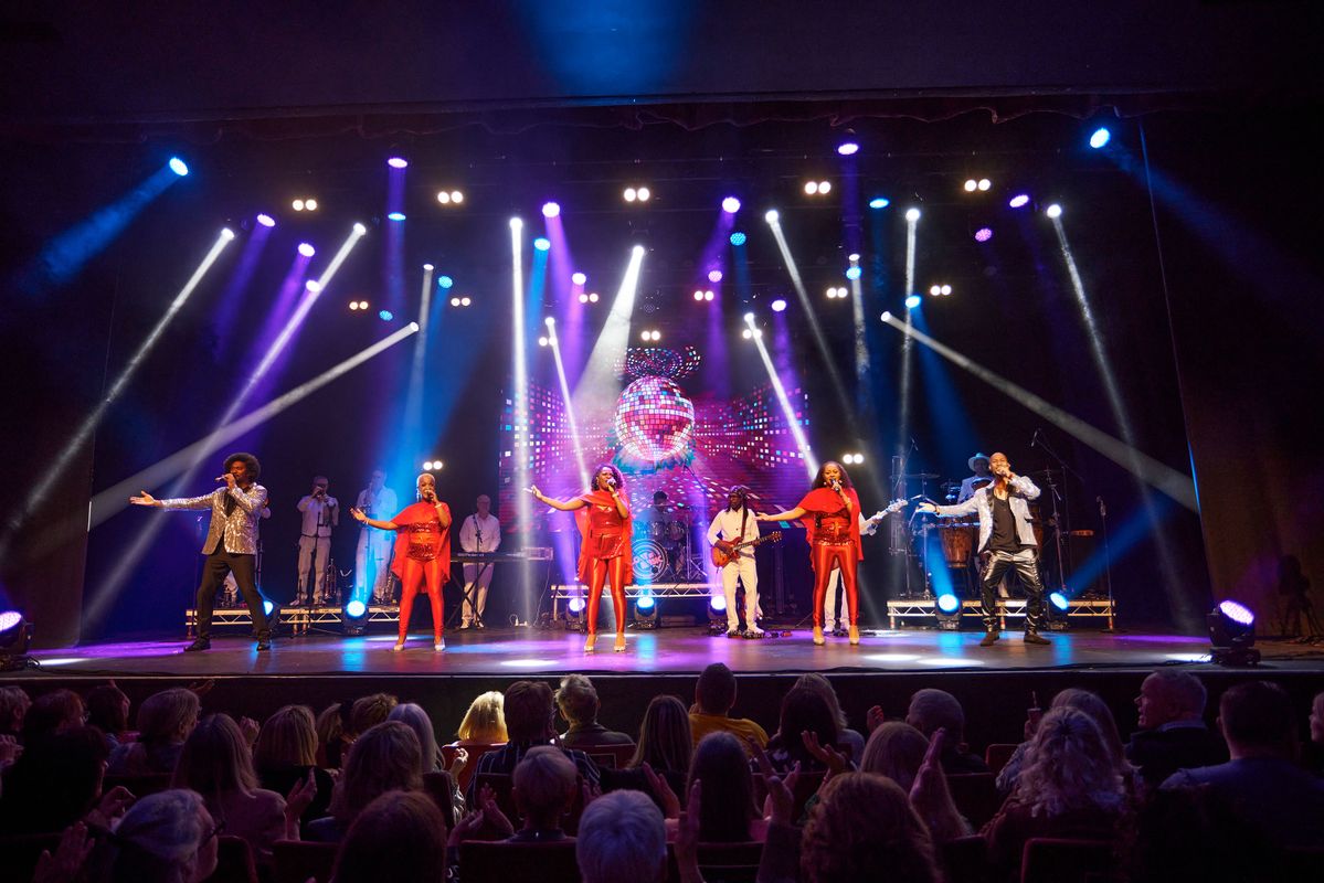 Multiple cast members wear sequined clothes with a disco ball image projected onto the screen behind.