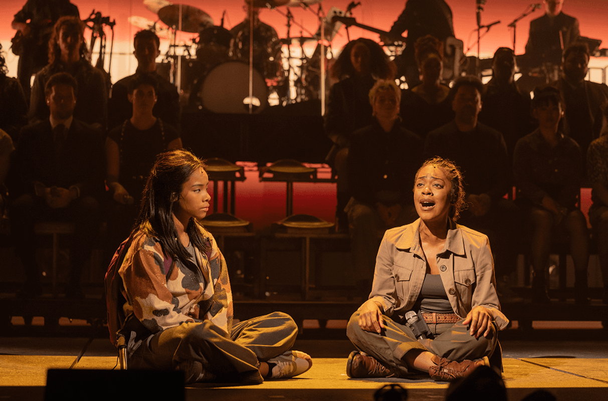 Two girls sat on the floor cross legged singing with a band behind them