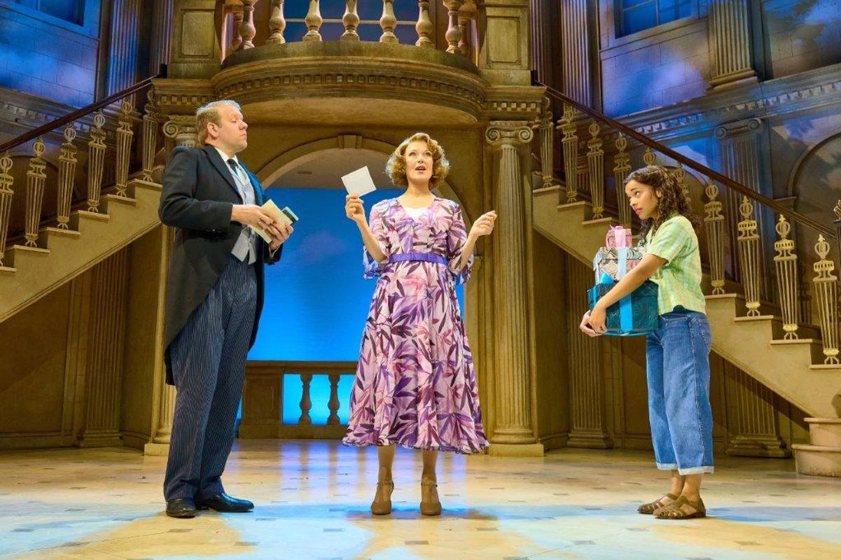 There are three people in a hall like setting, one is a butler, there is a woman stood in the middle holding a piece of paper and a younger girl beside her holding presents