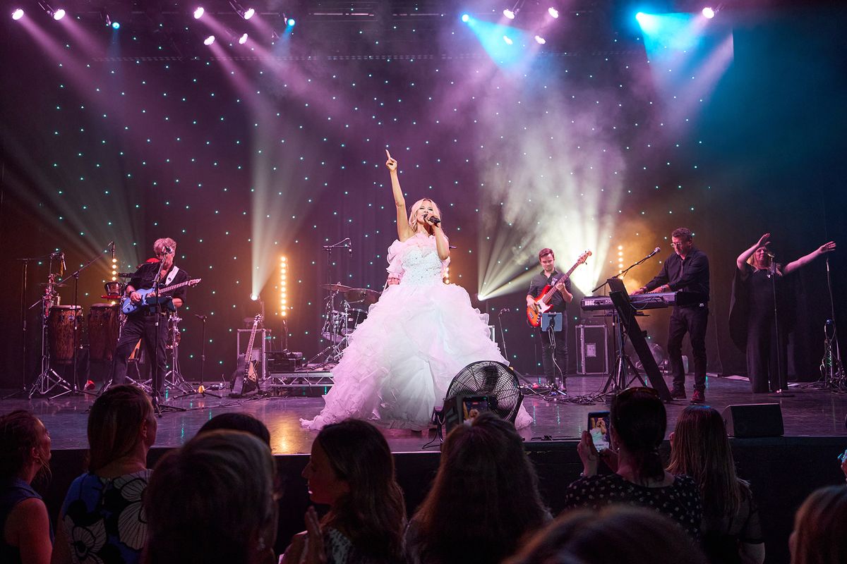 A lively concert scene shows a performer in a white, flowing gown on stage, passionately singing with one arm raised. The performer is accompanied by four musicians playing guitar, keyboard, and percussion. The stage is lit with colorful, bright lights and a backdrop of star-like dots. The audience is visible in the foreground, some with phones raised, capturing the moment.