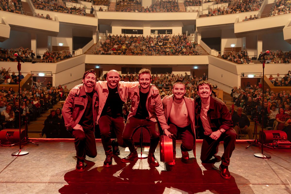 Five band members smile for the camera with the venue auditorium in the background