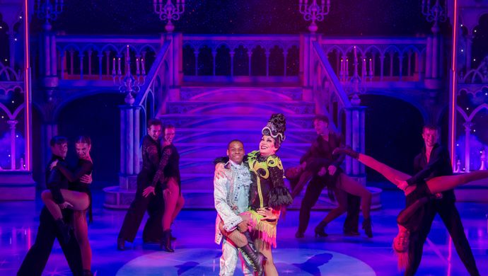 The wicked stepmother stands centre stage in a spotlight with their leg being held up by the prince.