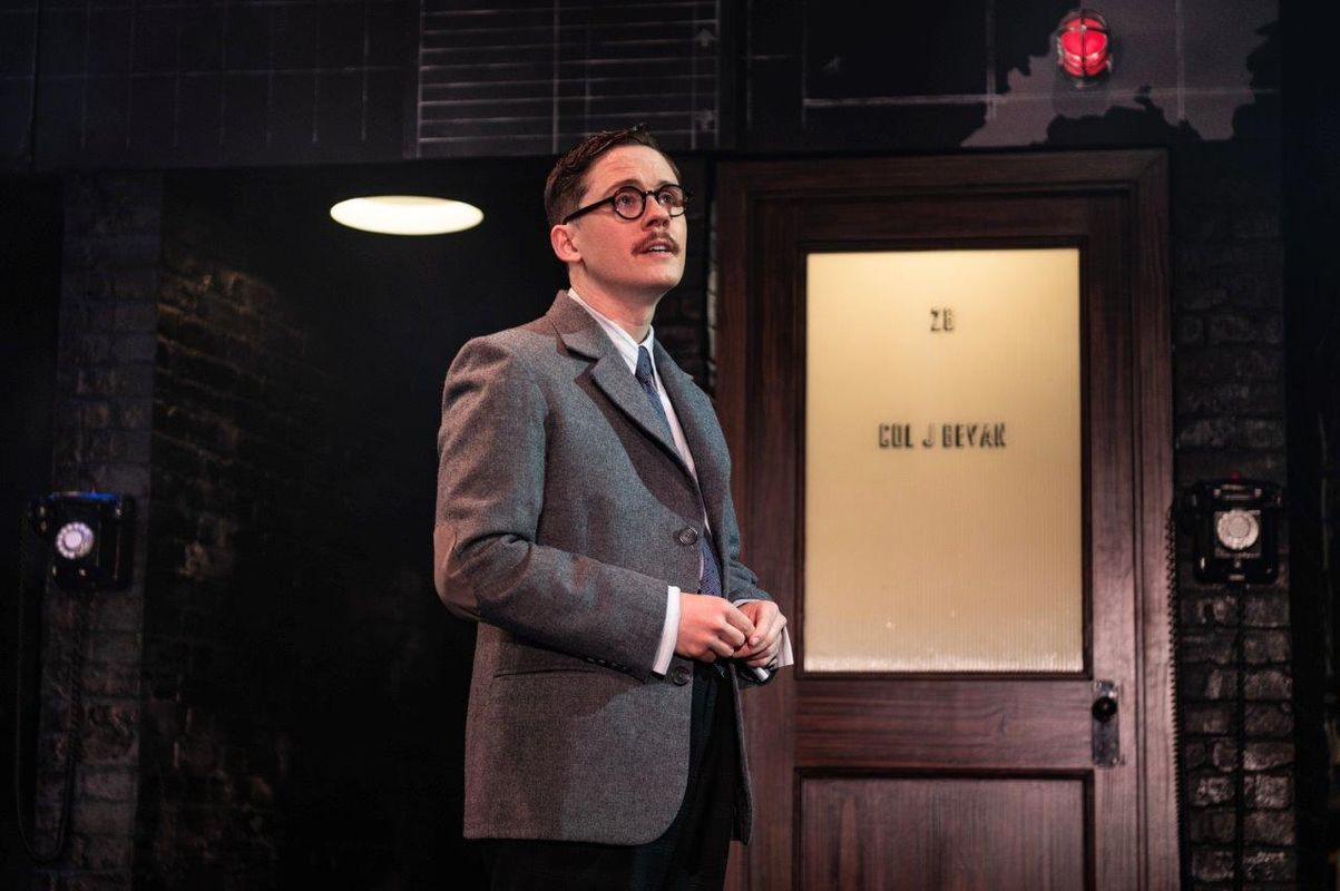 A man in a long a long coat and glasses. He looks to be in an office setting