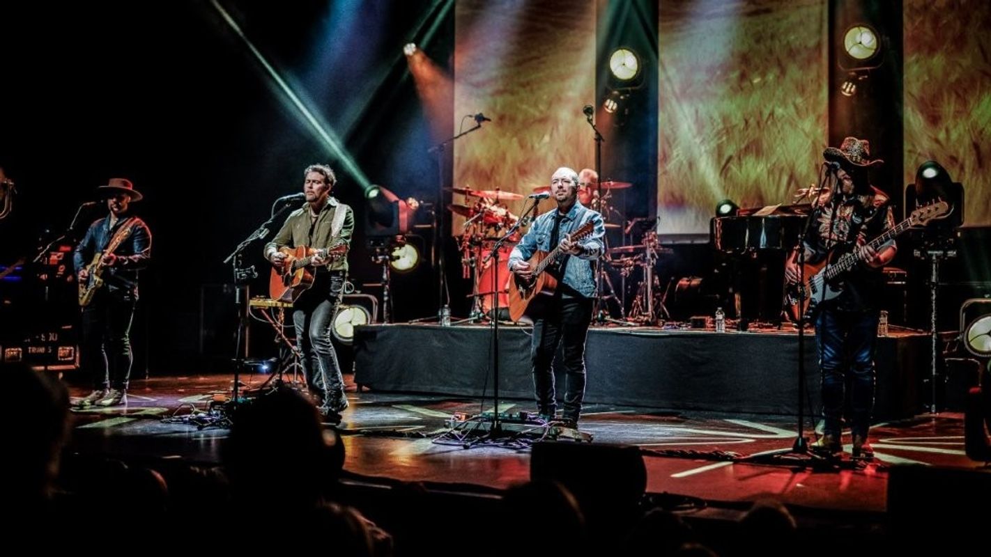 A band is performing on stage with guitars and a drum set. The backdrop is illuminated with stage lights, creating a lively atmosphere.