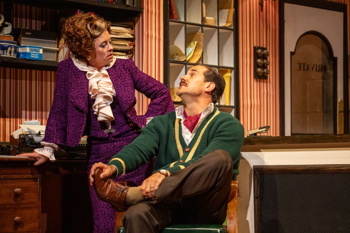A person in a purple suit leans on a desk next to someone sat on a chair.