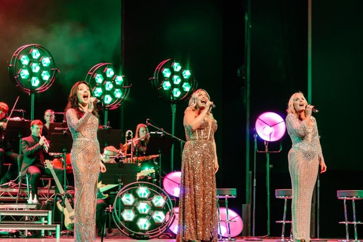Kerry Ellis, Louise Dearman and Rachel Tucker in sparkling gowns sing passionately on stage, lit by vibrant green and pink lights. A band accompanies them in the background.