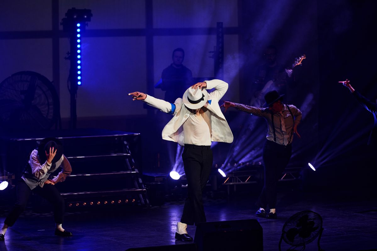 A dynamic stage performance featuring a dancer in a white suit and hat, striking a classic pose. Spotlight creates a dramatic, intense atmosphere.