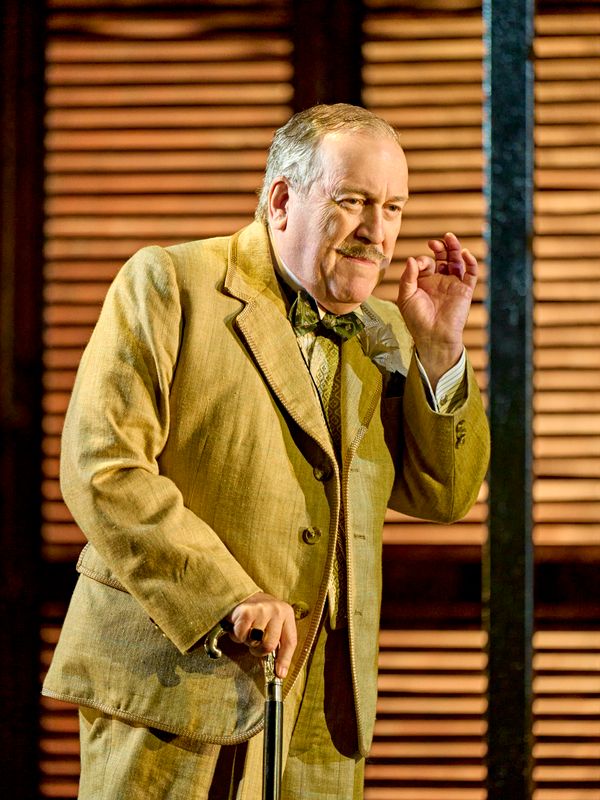 A person in a gold suit, with a walking stick, pretends to smoke a pipe.