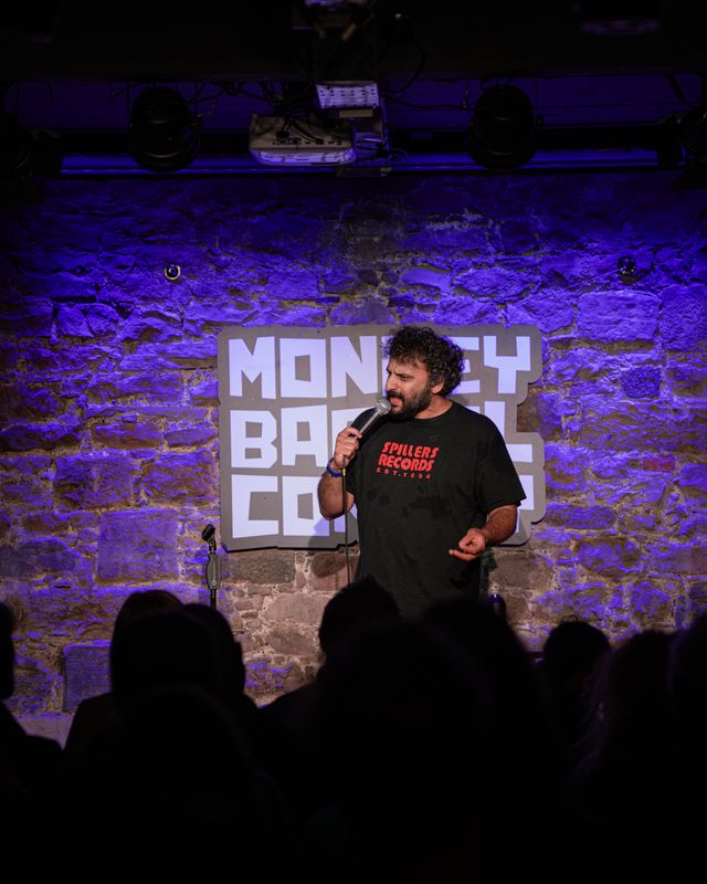 Nish stands onstage with a microphone wearing a Spillers Record t-shirt.